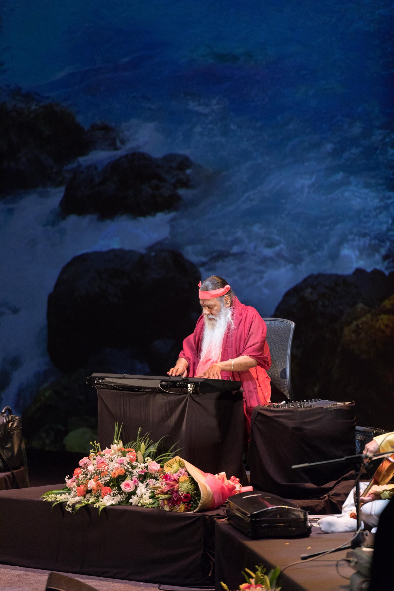 Sri Swamiji at keyboard — ocean waves backdrop